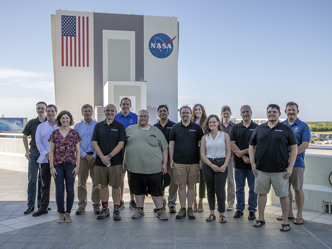 The ShadowCam team standing at Operations and Support Building II (OSB-II) at Kennedy Space Center (KSC) with the iconic NASA Vehicle Assembly Building (VAB) in the background.